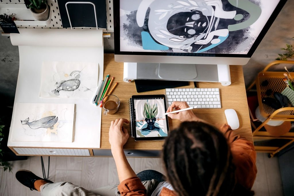 Top view of a designer working on digital artwork using a tablet and stylus at a creative desk setup with a monitor, sketches, and art supplies.
