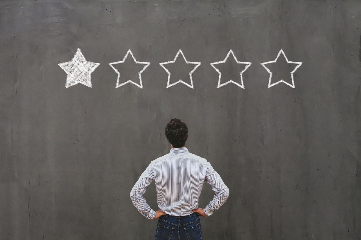 A person standing with hands on hips, looking at a chalkboard with a drawing of five stars, only one of which is filled in.