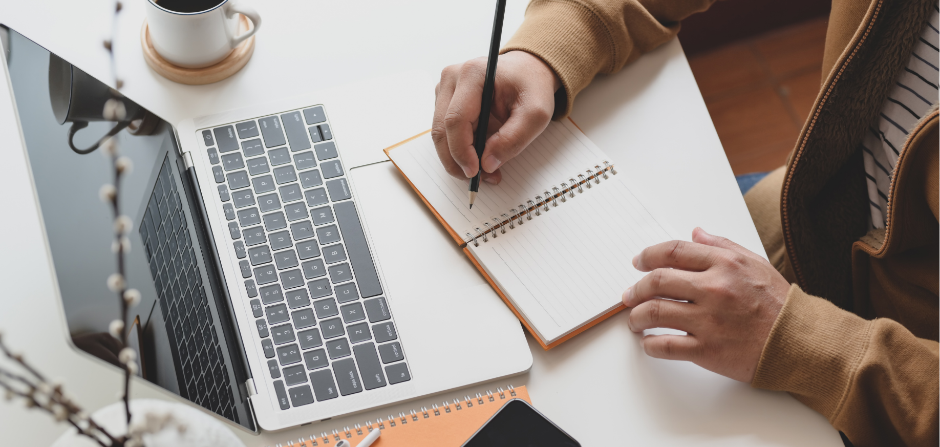 An overhead view of a person writing in a notebook next to an open laptop, on a white desk.