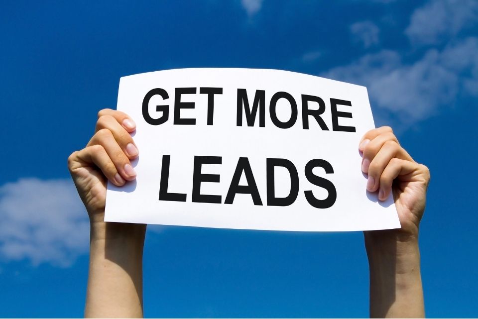Two hands holding up a white sign with the words 'GET MORE LEADS' in black text against a bright blue sky