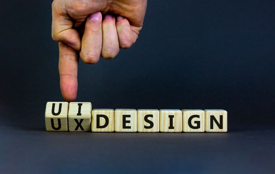 A hand pointing to wooden blocks that spell 'UI UX DESIGN' on a dark background.