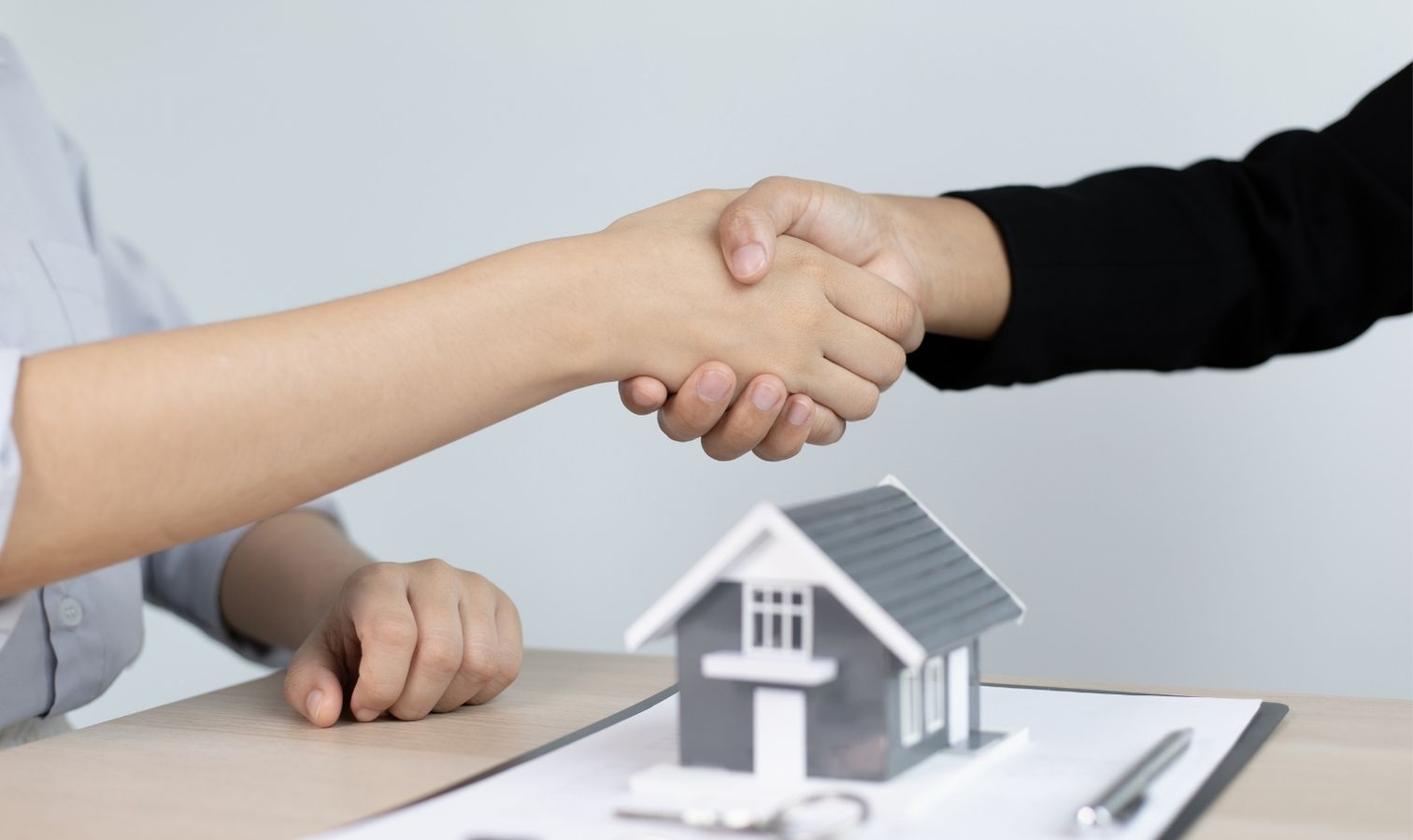 A real estate agent and a client shaking hands over a desk to finalize a property deal, with a miniature model house on the contract in the foreground