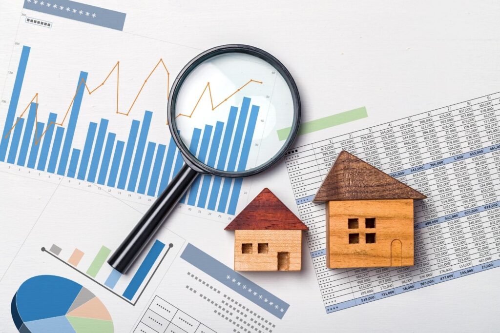A magnifying glass lies over financial charts and graphs, next to two small wooden model houses on a spreadsheet, symbolizing real estate market analysis and property valuation