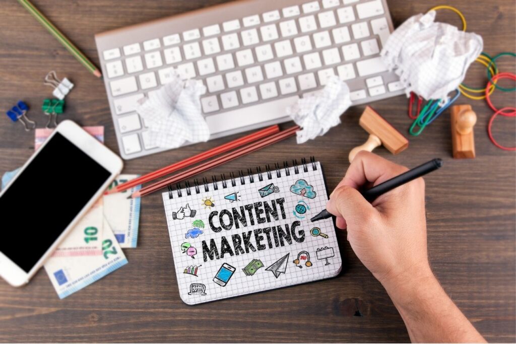 Person writing 'Content Marketing' on a notepad surrounded by a smartphone, money, keyboard, and office supplies, symbolizing creative digital strategy and planning