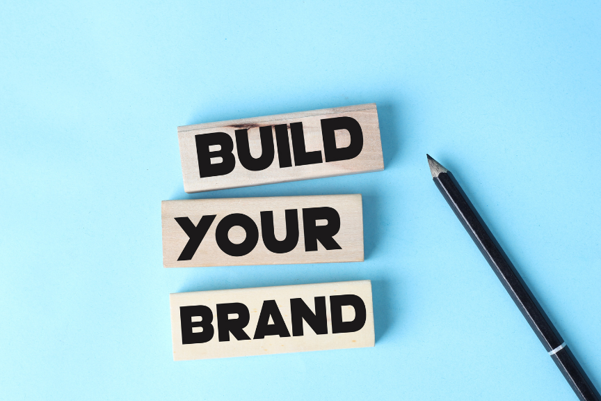 Three wooden blocks spelling 'BUILD YOUR BRAND' on a light blue background, next to a pencil