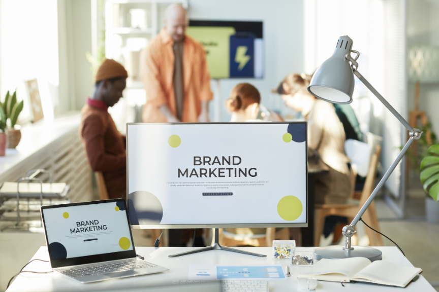 A computer monitor displaying 'BRAND MARKETING' with yellow and dark blue circles, with people working in the blurred backgroundD