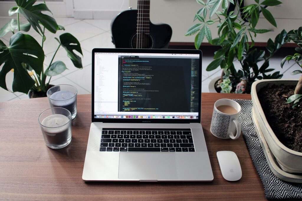 Laptop on a wooden desk displaying website code for Drupal development, surrounded by plants, candles, and a coffee cup