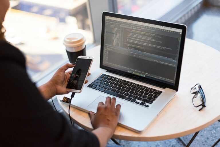 Person working on app development code on a laptop while holding a smartphone, seated at a round table with coffee and glasses nearby.