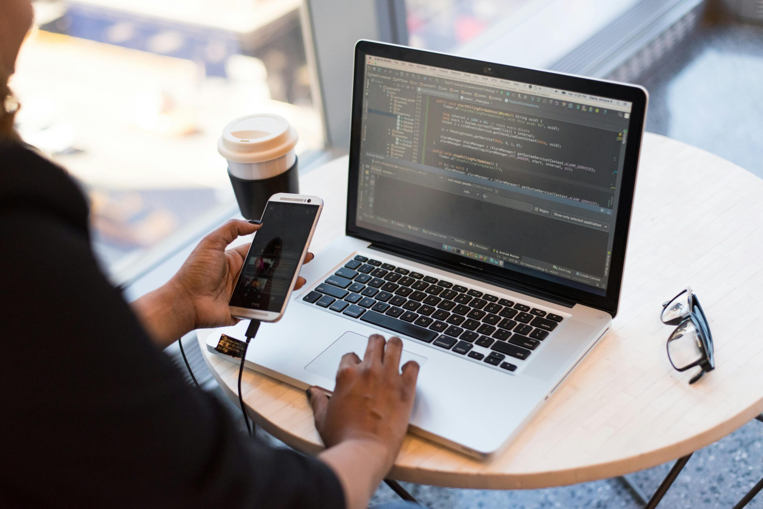 Person working on app development code on a laptop while holding a smartphone, seated at a round table with coffee and glasses nearby.