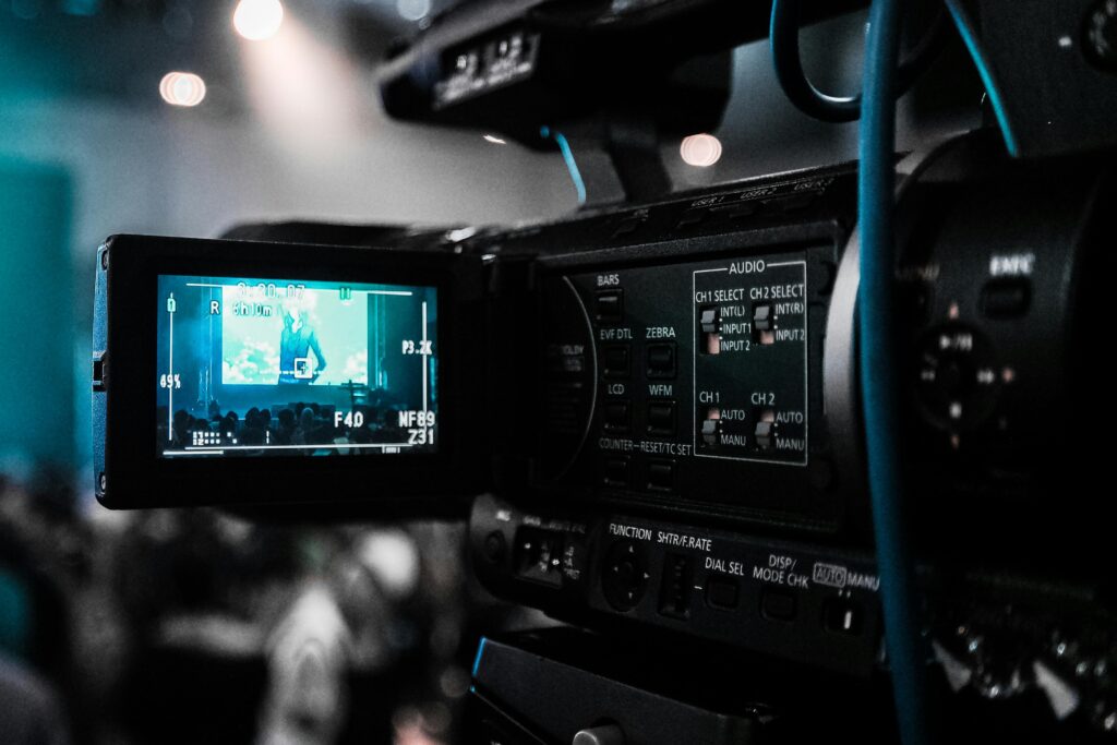 A close-up of a professional video camera capturing footage at a live event, with the on-screen monitor displaying the scene being recorded.