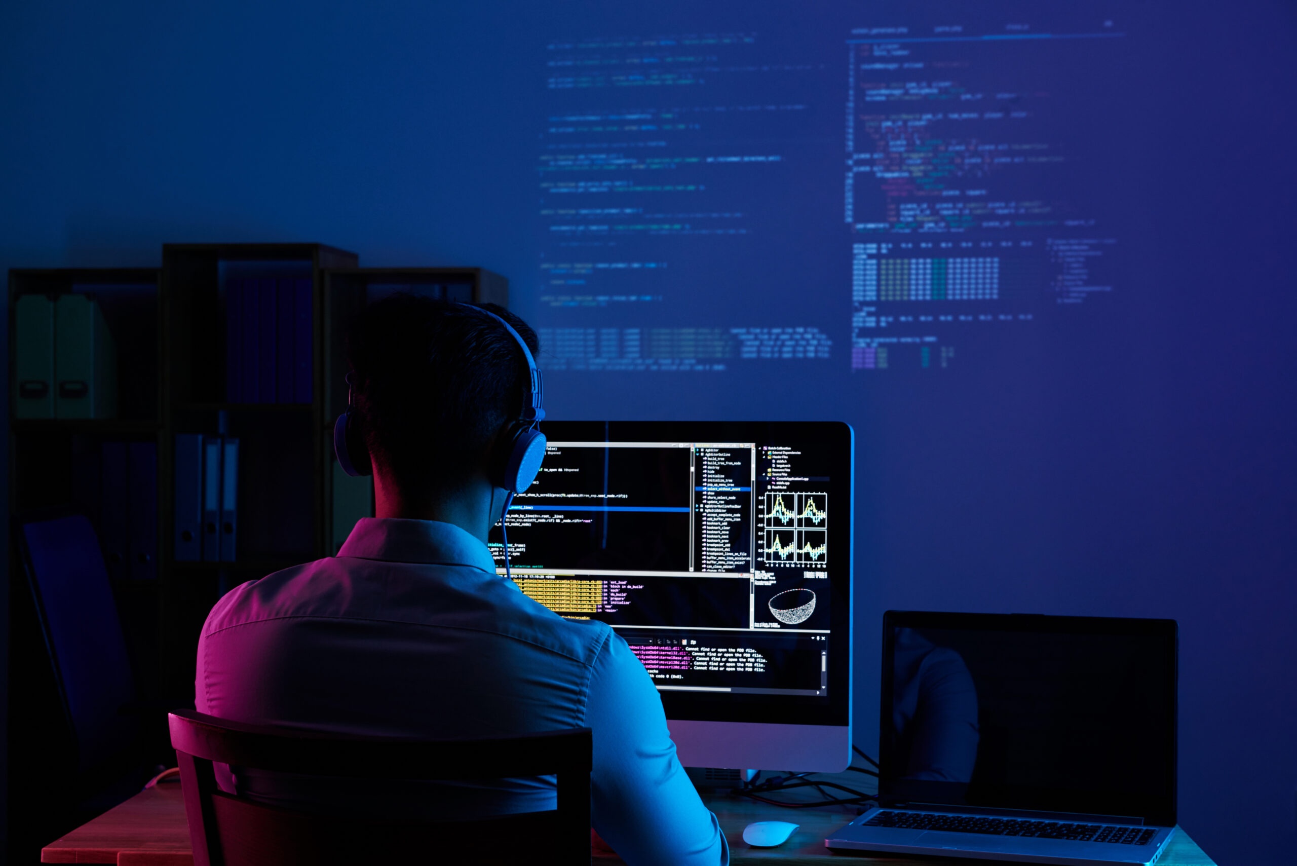 A developer wearing headphones, seen from behind, intently writing code on multiple computer screens in a dark room