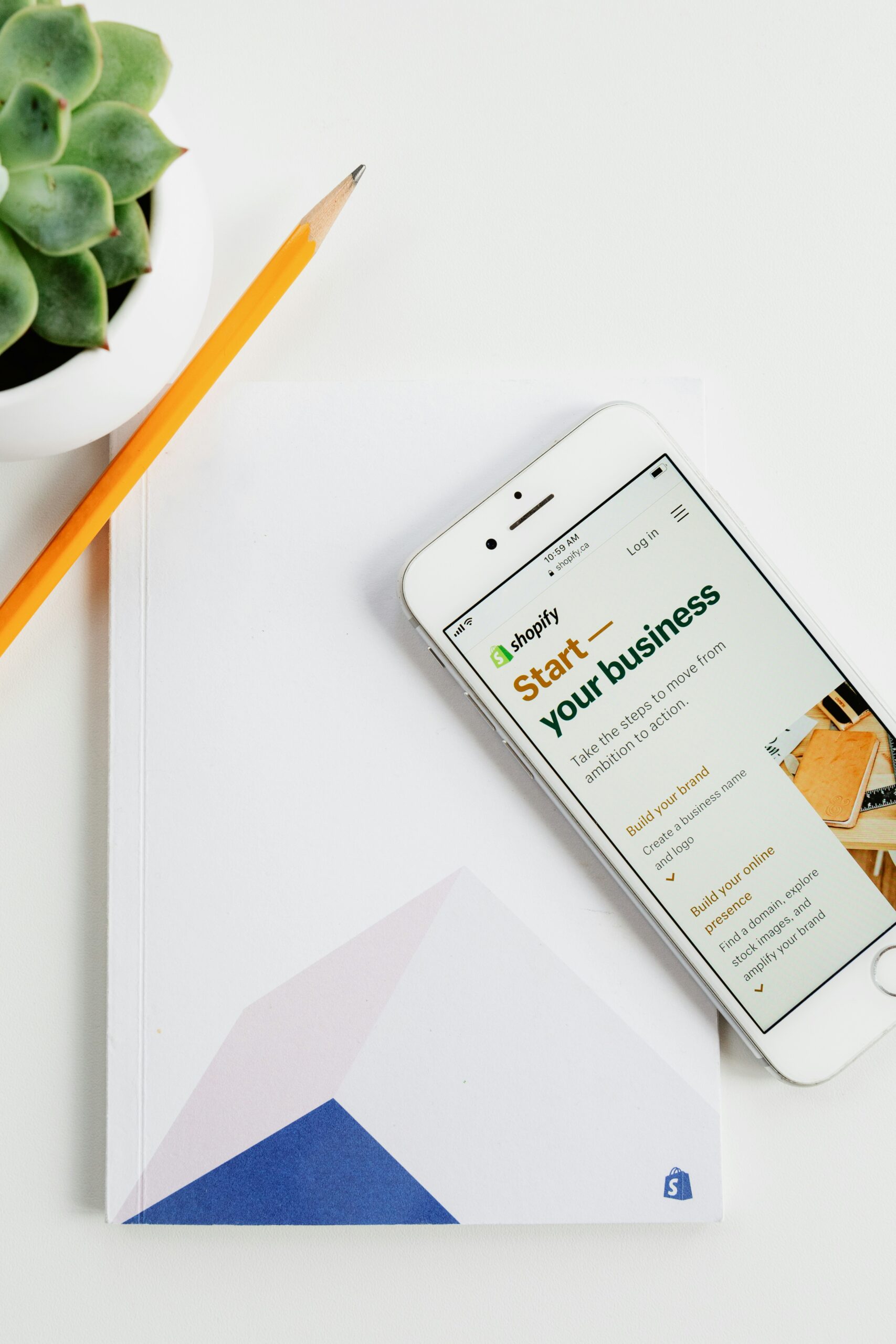 Smartphone with Shopify website open on a notebook beside a pencil and plant
