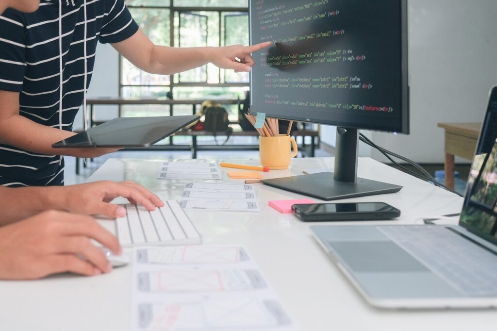 Developers collaborating on Magento web development project, reviewing code on a monitor and discussing website wireframes on a desk