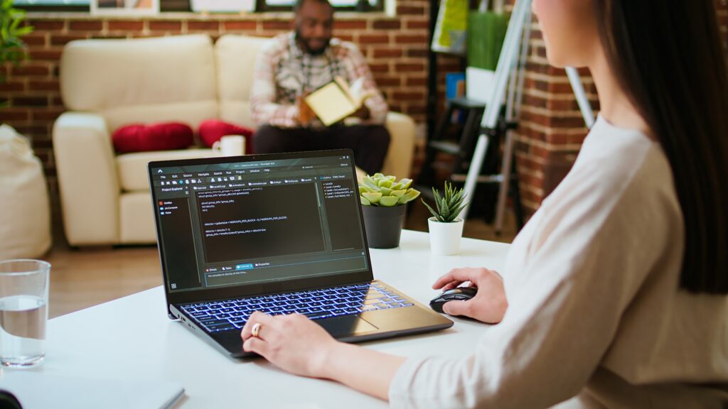 Programmer working on code at a laptop in a modern office with a colleague in the background.