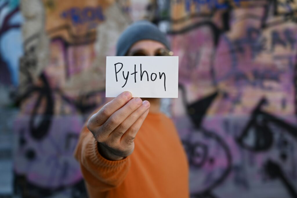 A person in a beanie holds up a small white card with "Python" written on it, standing in front of a colorful graffiti wall