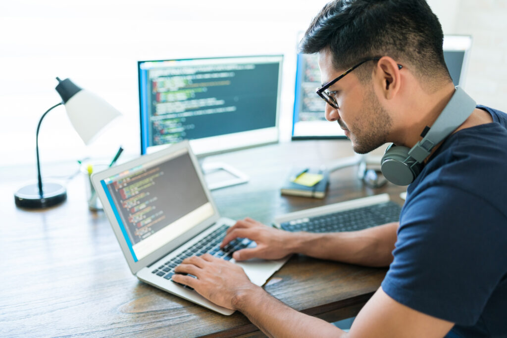 A focused male developer with glasses and headphones writing code on a laptop, with two additional monitors displaying code in the background